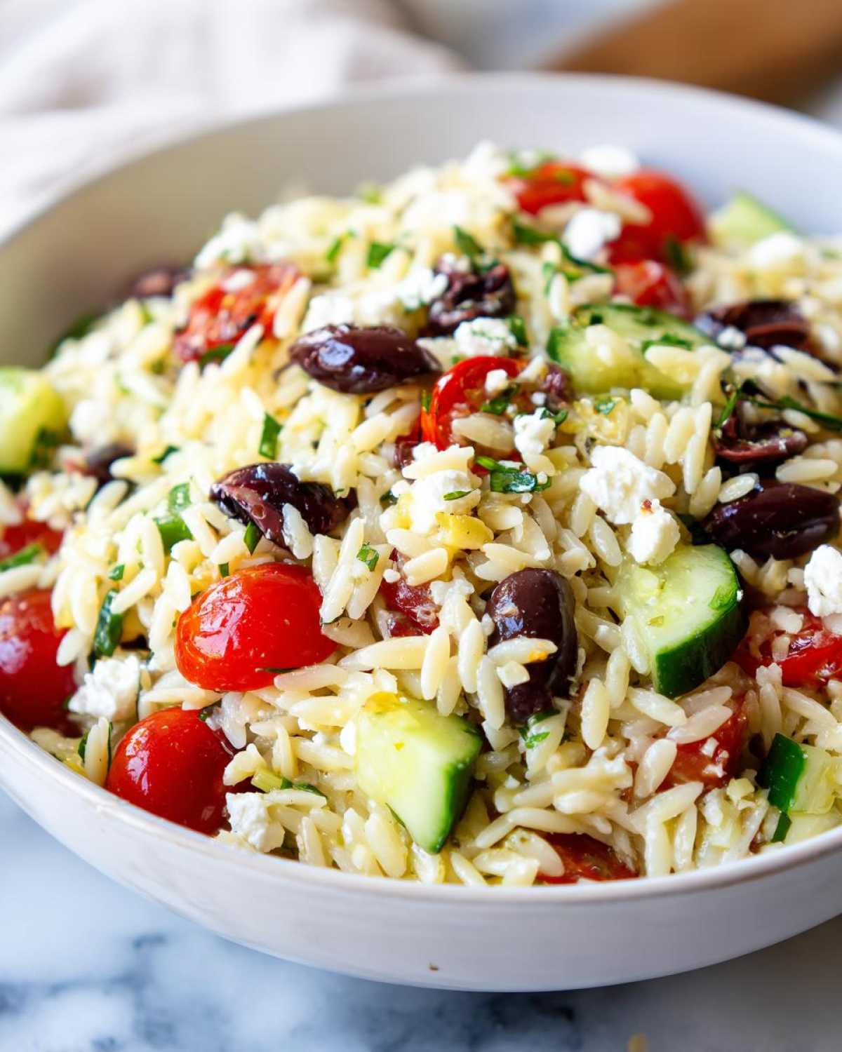 A close-up view of a bowl filled with Mediterranean Orzo Salad with Feta, featuring orzo pasta, cherry tomatoes, cucumbers, olives, and feta cheese.