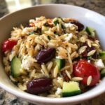 Close-up of a white bowl filled with Mediterranean Orzo Salad with Feta, featuring orzo pasta, Kalamata olives, cucumber, and tomatoes.