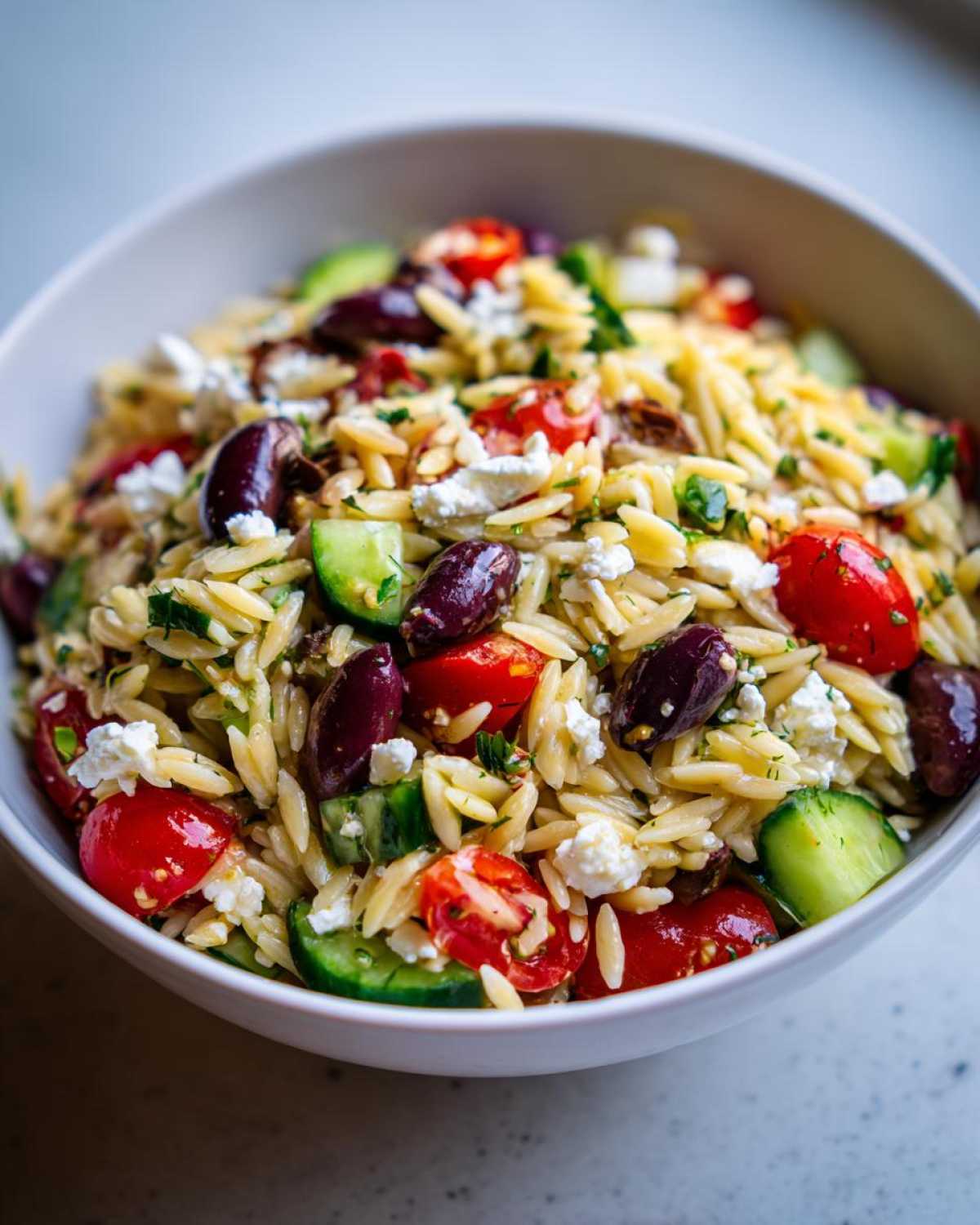 A close-up view of a white bowl filled with Mediterranean Orzo Salad with Feta, featuring orzo pasta, cherry tomatoes, cucumbers, and Kalamata olives.