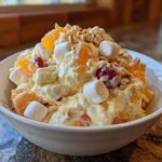 A close-up of a white bowl filled with creamy Memorial Day Ambrosia Salad, featuring mandarin oranges, pineapple, cherries, marshmallows, and chopped nuts.