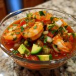 A close-up of vibrant Mexican shrimp cocktail featuring plump shrimp, avocado chunks, onion, and cilantro in a tomato-based sauce.