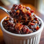 A spoonful of rich, dark Million Dollar Baked Beans topped with crispy bacon pieces being lifted from a white ramekin.