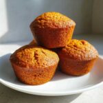 Three golden-brown, moist pumpkin spice muffins stacked slightly on a white plate, catching bright sunlight.
