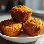 Three moist pumpkin spice muffins stacked on a small white plate, highlighted by natural sunlight.