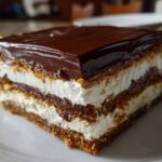 A close-up of a layered slice of No-Bake Chocolate Eclair Cake featuring graham crackers, cream filling, and chocolate glaze.