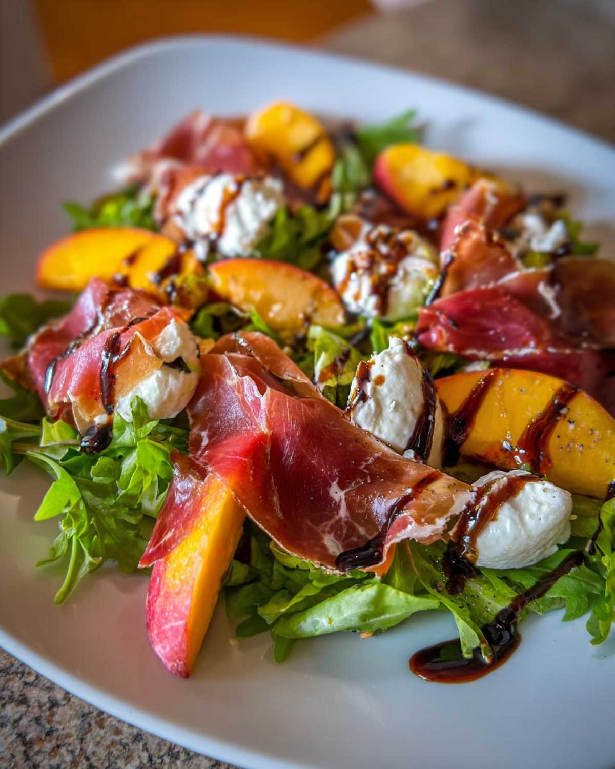 A vibrant Peach & Burrata Salad with Prosciutto served on a white plate, drizzled with balsamic glaze over arugula.