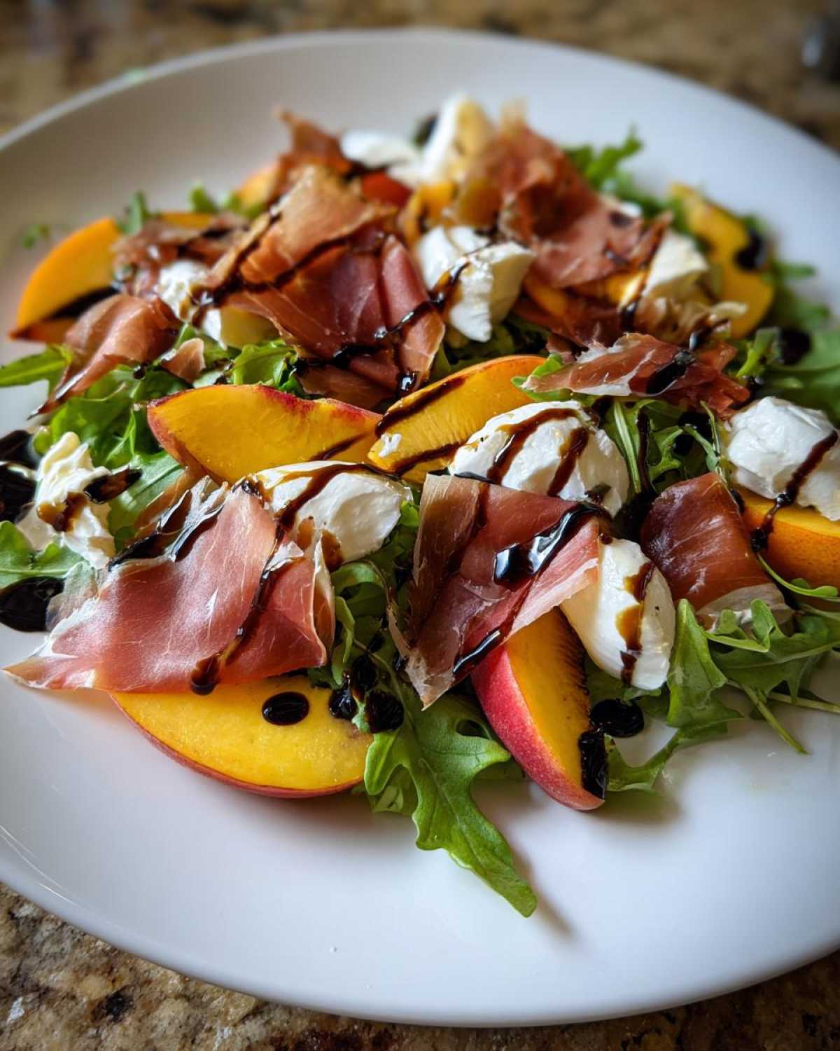 Close-up of a vibrant Peach & Burrata Salad with Prosciutto, arugula, and balsamic glaze on a white plate.