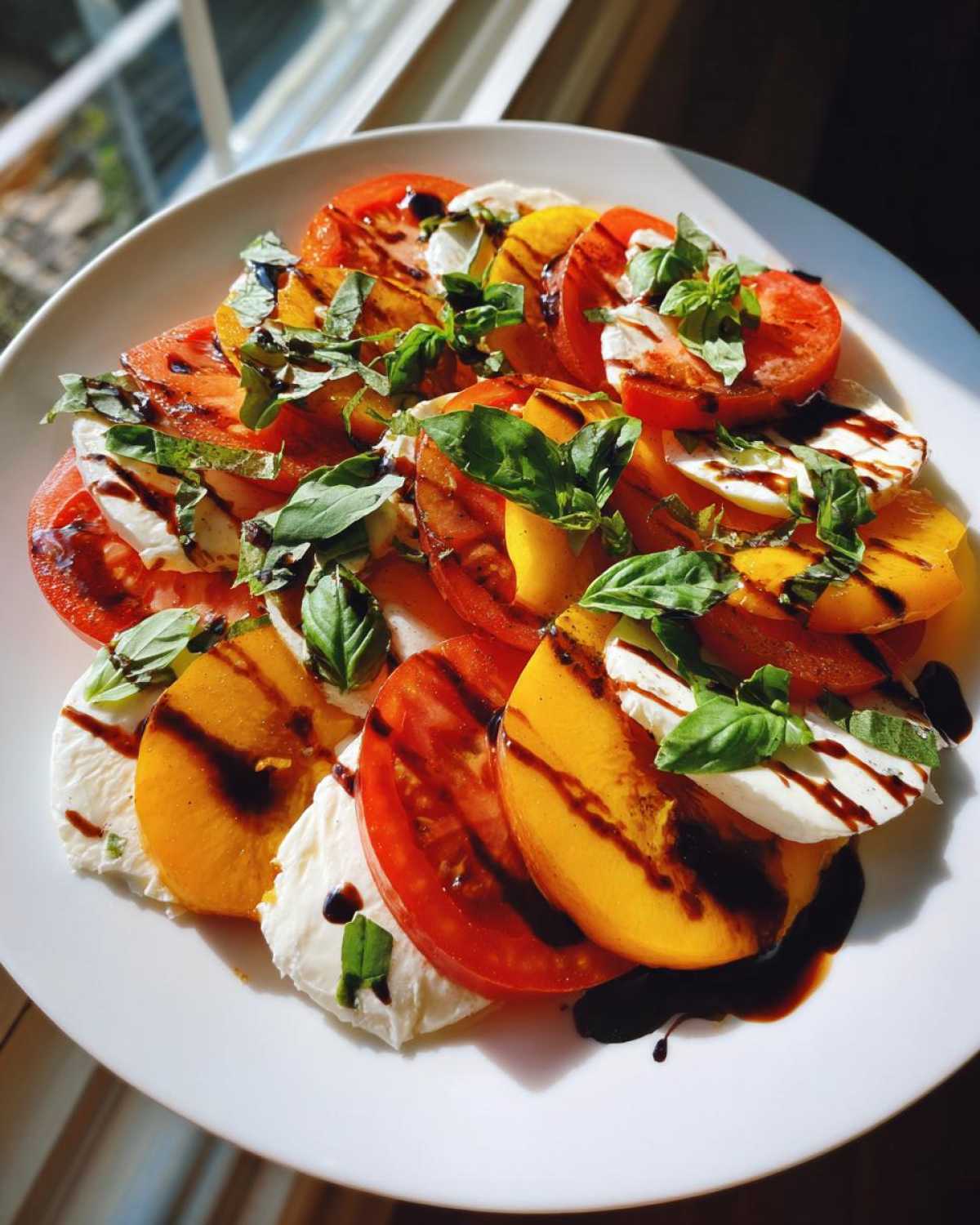 A plate featuring slices of tomato, fresh mozzarella, and yellow peaches arranged in a Peach Caprese Salad, drizzled with balsamic glaze and topped with basil.
