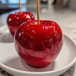 Close-up of a perfectly dipped, shiny red candy apple on a white plate, ready to eat.