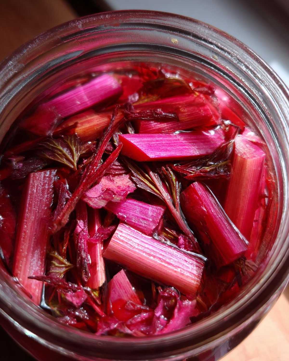 Close-up overhead view of bright pink pickled rhubarb stalks and red shiso leaves soaking in liquid inside a glass jar.