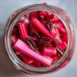 Overhead view of bright pink Pickled Rhubarb with Red Shiso pieces packed tightly in a clear glass jar.