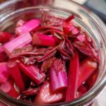 Close-up view inside a glass jar showing vibrant pink and red stalks of Pickled Rhubarb with Red Shiso submerged in liquid.