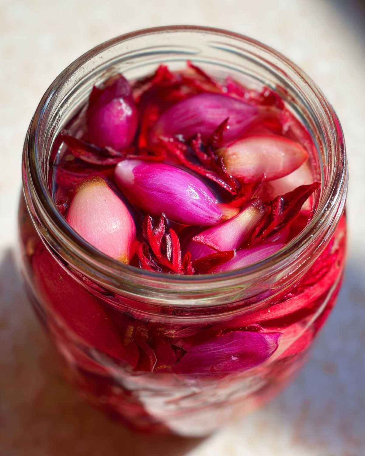 Close-up overhead view of a glass jar filled with bright pink pickled rhubarb pieces and red shiso leaves soaking in liquid.