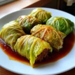 Close-up of six savory cabbage rolls served on a white plate swimming in a rich, dark sauce.