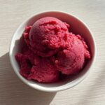 Three scoops of vibrant, deeply colored Raspberry Sorbet served in a small white bowl on a light wooden surface.