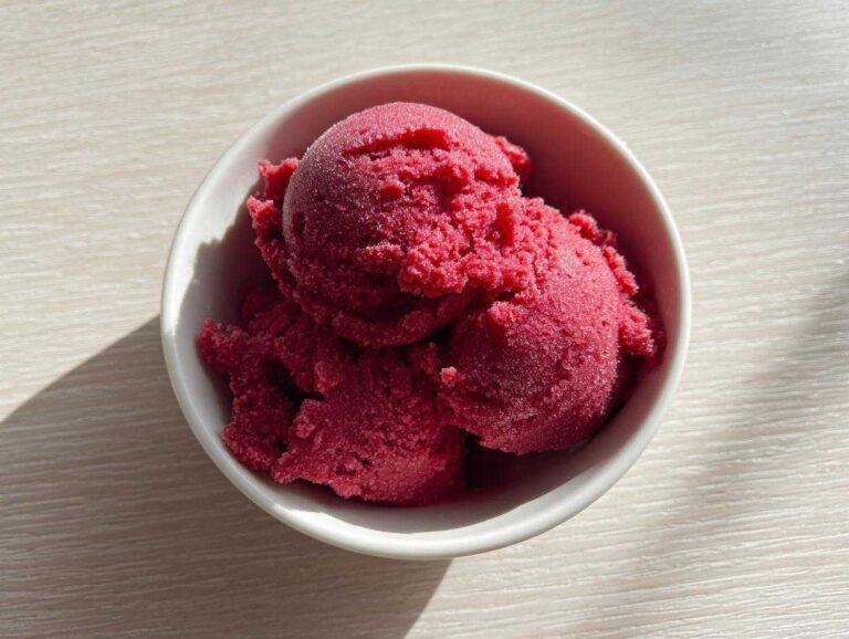 Three scoops of vibrant, deeply colored Raspberry Sorbet served in a small white bowl on a light wooden surface.