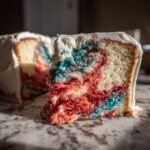 Close-up of a slice showing the vibrant red and blue marbled interior of the Red, White & Blue Marble Memorial Day Cake, covered in white frosting.