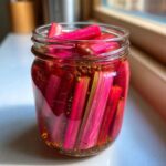 Bright pink rhubarb stalks packed tightly in a glass jar with pickling liquid and black peppercorns for Refrigerator Rhubarb Pickles.