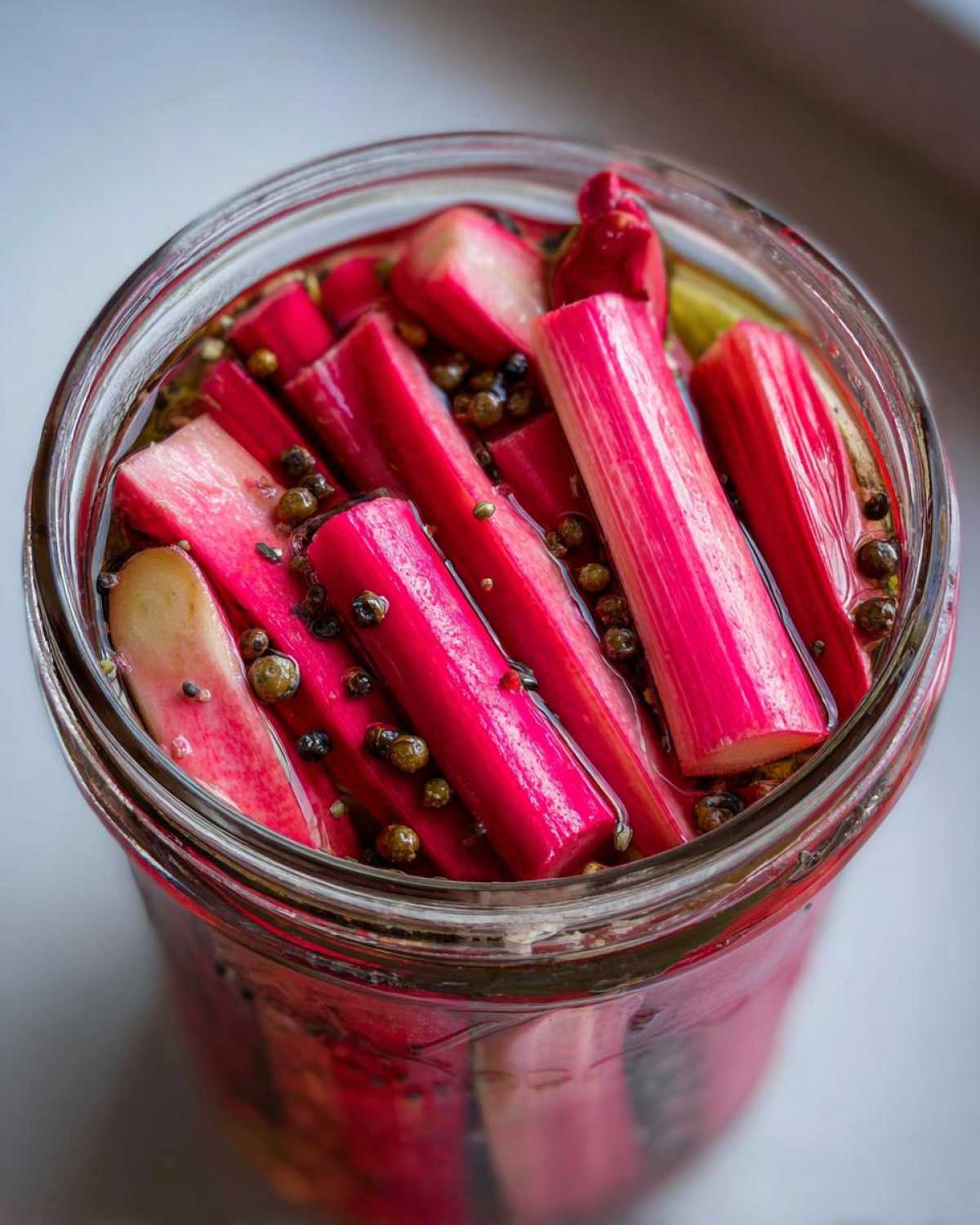 Close-up overhead view of bright pink Refrigerator Rhubarb Pickles with black peppercorns in a glass jar.