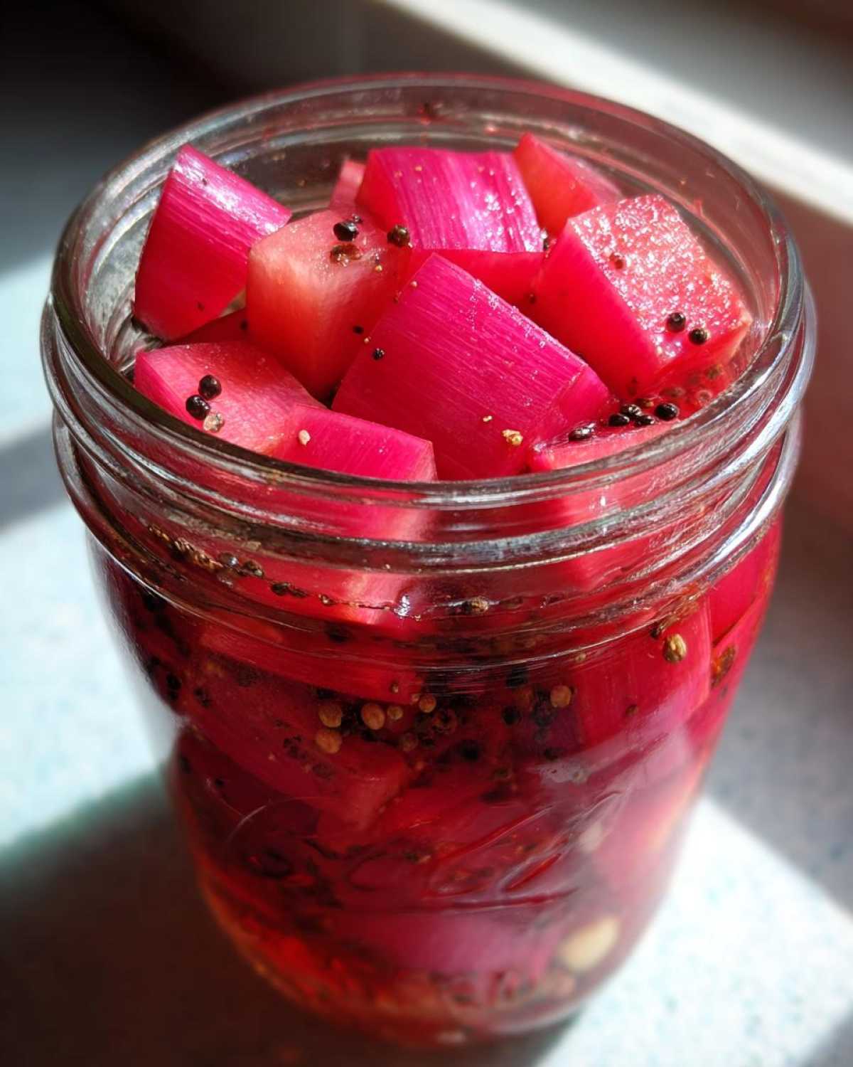 Close-up of bright pink Refrigerator Rhubarb Pickles chunks packed tightly in a glass jar with black peppercorns.