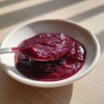A spoonful of vibrant, deep red Rhubarb and Beetroot Ketchup being lifted from a small white bowl.