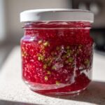 Close-up of a jar filled with bright red Rhubarb and Elderflower Jam, showing suspended fruit pieces and elderflower blossoms.