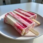 Two frozen Rhubarb and Greek Yogurt Popsicles with pink and white stripes resting on a white plate.