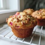 A close-up of a freshly baked Rhubarb and Pecan Muffin with a crunchy pecan topping cooling on a white wire rack.