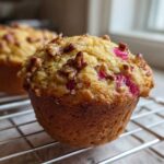 A close-up of a golden-brown Rhubarb and Pecan Muffins cooling on a wire rack, showing bright pink rhubarb pieces.