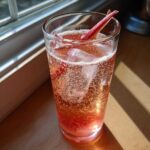A tall glass filled with a fizzy, pink Rhubarb Cocktail with Ginger Beer, ice, and a rhubarb stalk garnish.