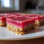 Close-up of bright pink Rhubarb Curd Shortbread squares with a crumbly base, served on a white plate.