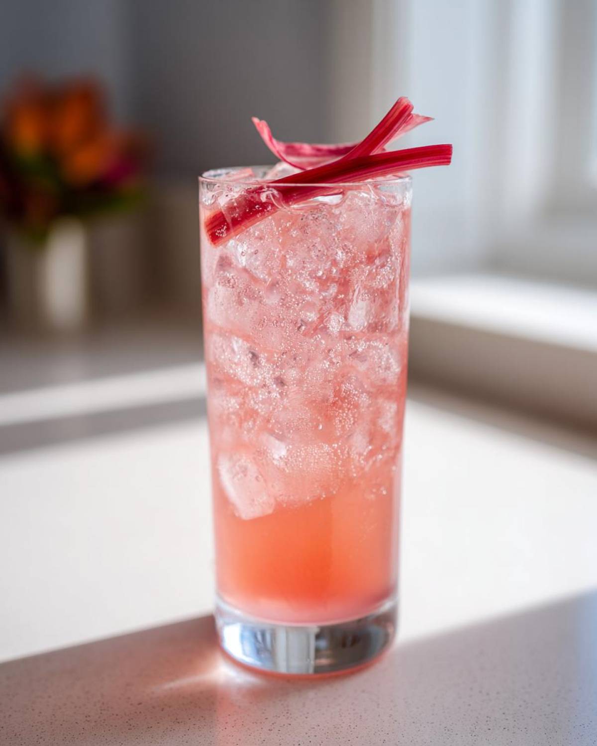 A tall glass filled with a pink, bubbly Rhubarb and Elderflower Gin Fizz cocktail over ice, garnished with rhubarb strips.