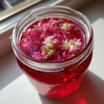 Close-up of pink rhubarb-infused liquid with elderflower blossoms floating on top in a glass jar, part of making Rhubarb and Elderflower Jam.