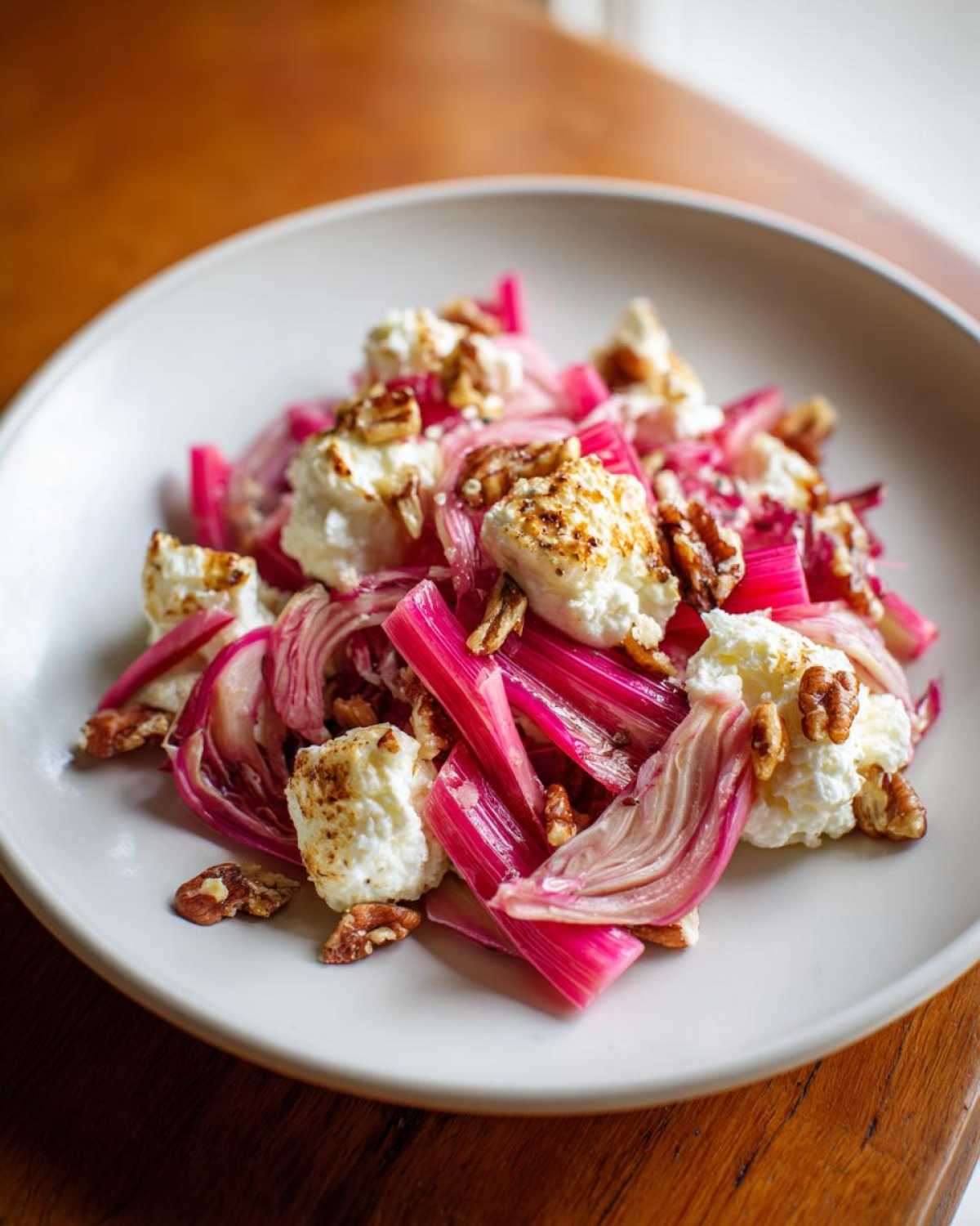 A close-up of a Rhubarb Fennel Salad featuring bright pink rhubarb, roasted ricotta dollops, and pecans on a white plate.