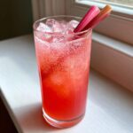 A tall glass filled with a bright pink, fizzy Rhubarb Fizz Cocktail, garnished with two rhubarb stalks.