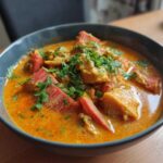 A close-up of a rich, orange Rhubarb-Lentil Curry served in a dark bowl, topped with fresh parsley.