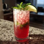 A tall glass of bright pink Rhubarb Mojito with fresh mint and a lime wedge garnish, sitting on a granite counter.