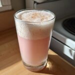 A tall glass of pink Rhubarb Rose Ramos Gin Fizz topped with thick white foam and a sprinkle of spice.