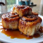 Four gooey Rhubarb Sticky Buns with Cardamom stacked on a white plate, dripping with rich caramel glaze.