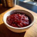 A small white bowl filled with thick, glossy, deep red Rhubarb-Wine BBQ Sauce, sitting on a wooden surface.