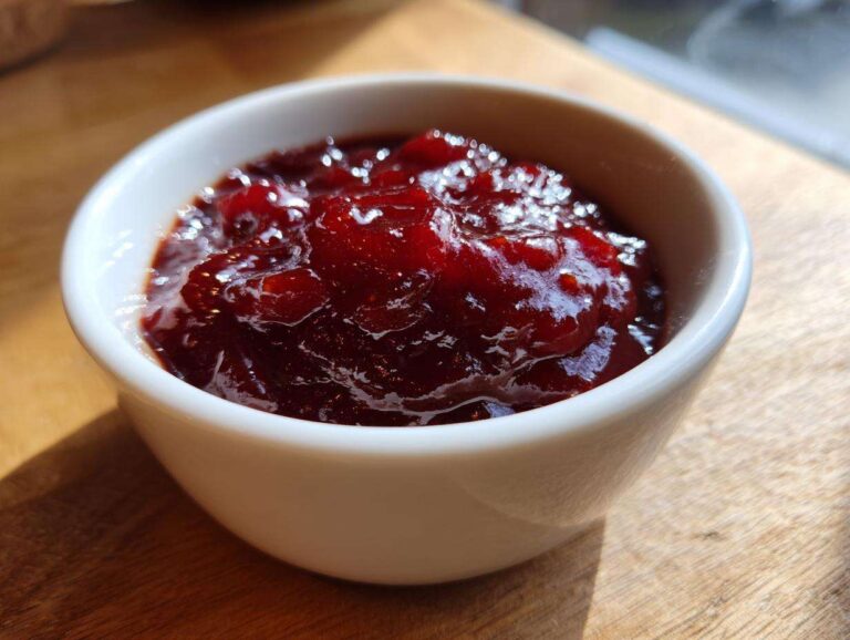 Close-up of thick, glossy, dark red Rhubarb-Wine BBQ Sauce served in a small white bowl on a wooden surface.