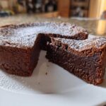 A rich, dark flourless chocolate torte with a slice cut out, dusted lightly with powdered sugar.