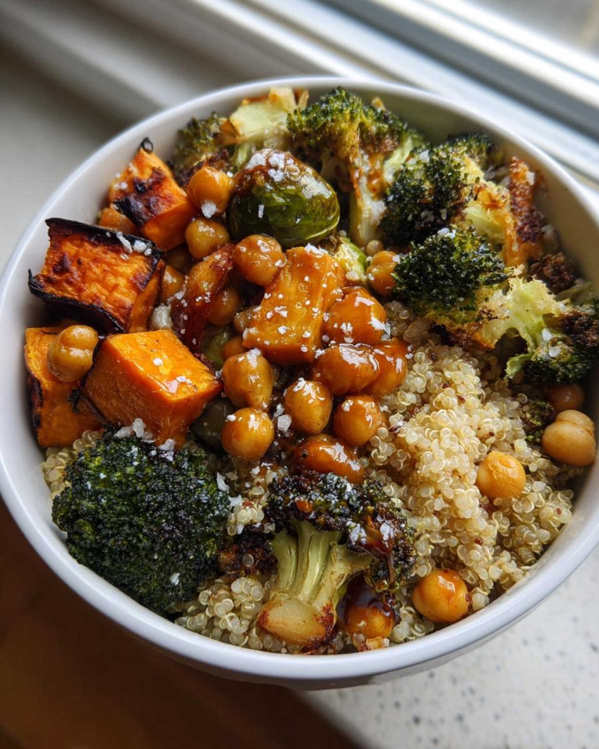 Close-up of a white bowl filled with Roasted Vegetable Quinoa Salad featuring roasted sweet potatoes, broccoli, chickpeas, and Brussels sprouts and a glaze.