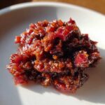 A close-up, glossy mound of deep red Savory Rhubarb Chutney with Dates served on a white plate.