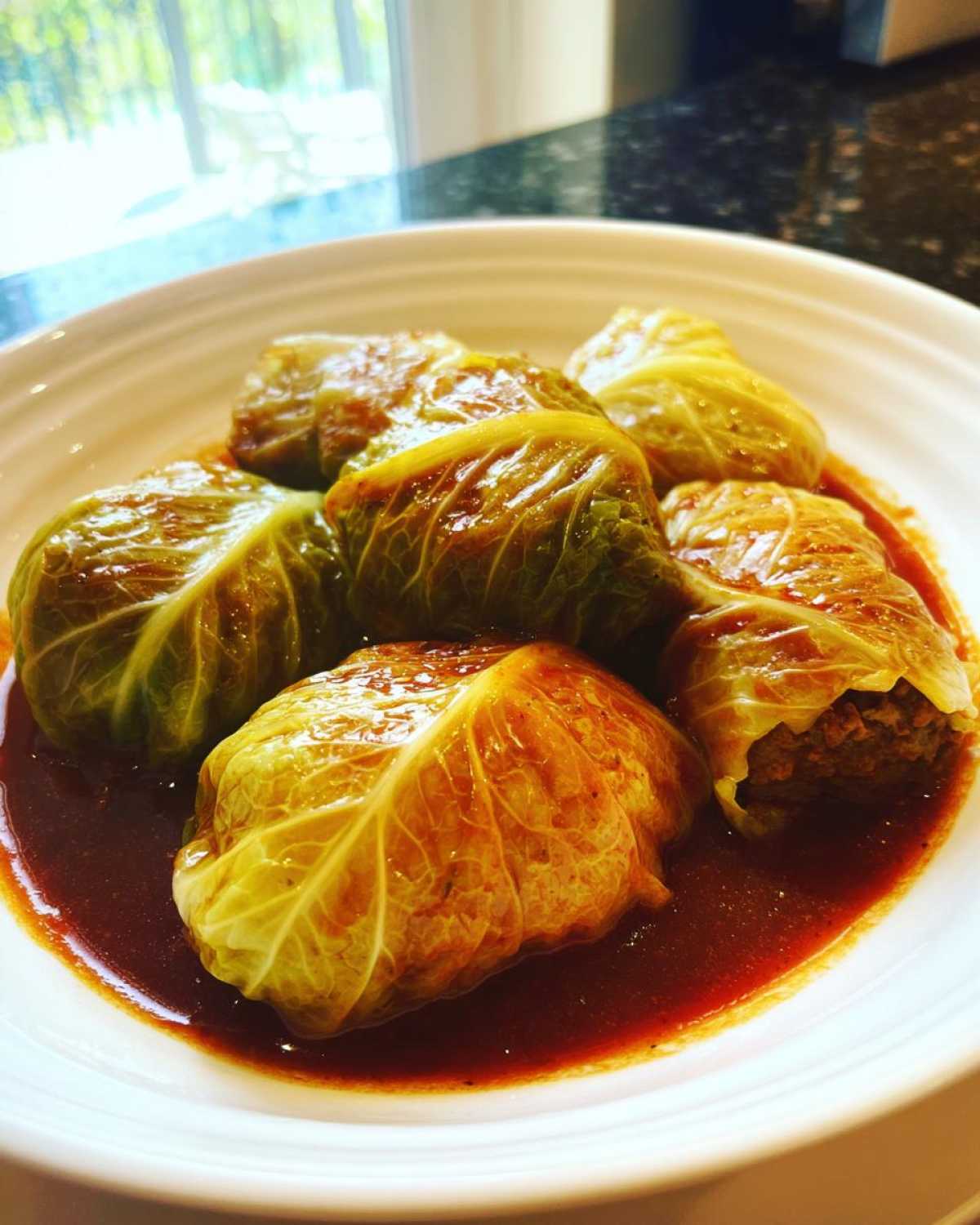 A white bowl filled with several savory cabbage rolls simmering in a rich, dark red sauce.