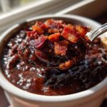 A spoonful of rich, dark Million Dollar Baked Beans topped with crispy bacon bits being lifted from a white ramekin.
