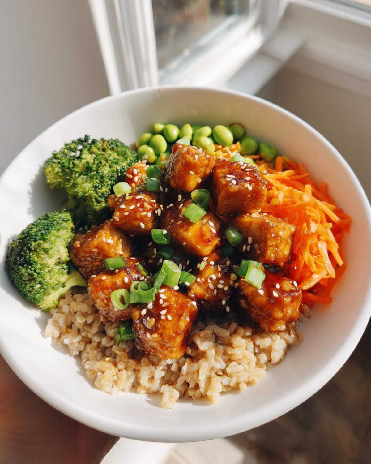 A white bowl filled with Sesame Ginger Tofu Bowls, featuring glazed tofu, brown rice, broccoli, shredded carrots, and edamame.