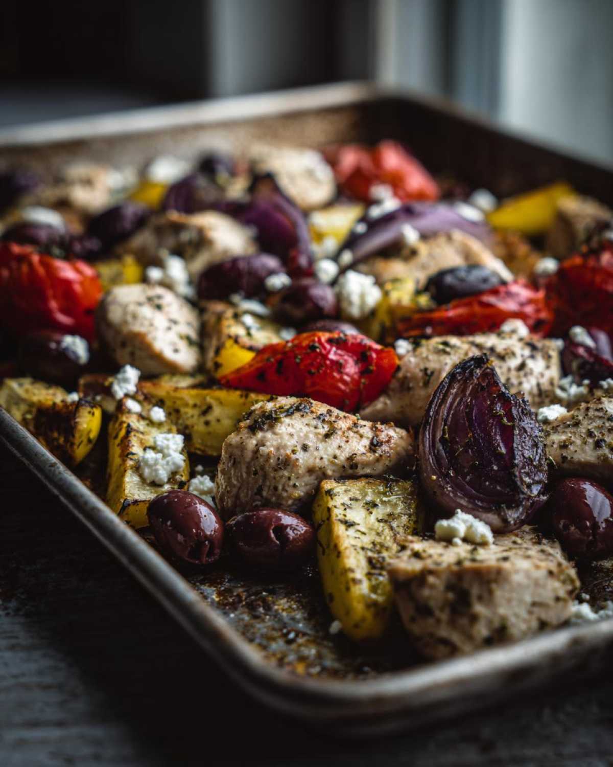 Close-up of Sheet Pan Greek Chicken & Veggies with roasted peppers, onions, olives, and feta on a metal tray.