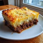 A square slice of rich cottage pie featuring a thick layer of browned ground meat topped with golden, crispy mashed potatoes.