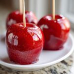 Three shiny, deep red candy apple treats on sticks, resting on a white plate, with one in the foreground sparkling.